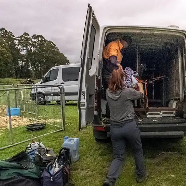 The rear door of a mercedes van is open. A woman is taking some shearing equipment from a man standing inside the van.