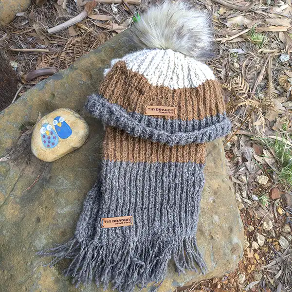 Tri-colour (white, brown and charcoal) beanie and scarf with fringe are laid flat on a large rock.