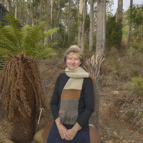 An older woman is weraing a tri-colour (white, brown, charcoal) beanie and matching scarf in front of a nautral bush setting.