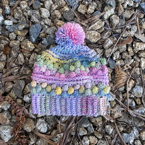 A multi-coloured pink and blue hand-crocheted beanie is lying flat on a bed of gravel. It has a pom-pom on the top.