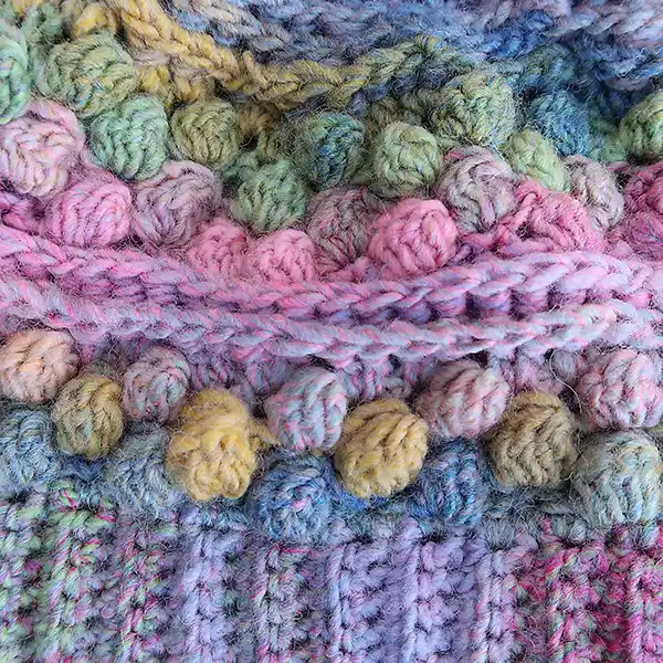 A multi-coloured pink and blue hand-crocheted beanie is lying flat on a bed of gravel. It has a pom-pom on the top. This image shows a close up of the pattern.