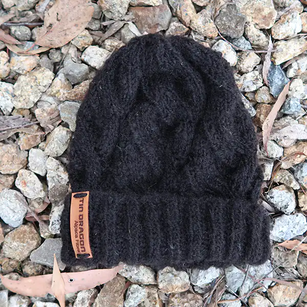 A black kintted alpaca beanie is photographed from above, as it lays flat on small grey-coloured stones.