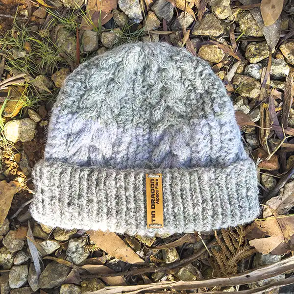 A hand-knittted beanie styled with cable-knit is displayed on a gravel backgound