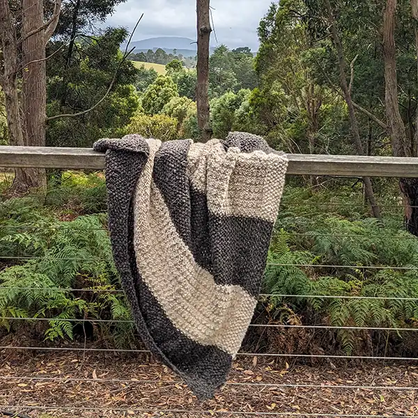 Pure alpaca blanket in charcoal and cream colour is spread lossley over a fence with green bushland and mountains in the background