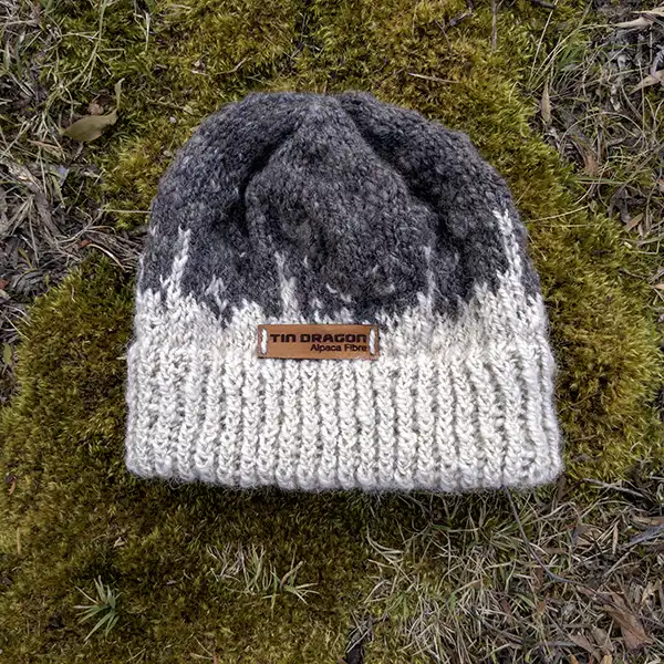 Awhite and chacoal coloured alpaca beanie is sitting on green moss