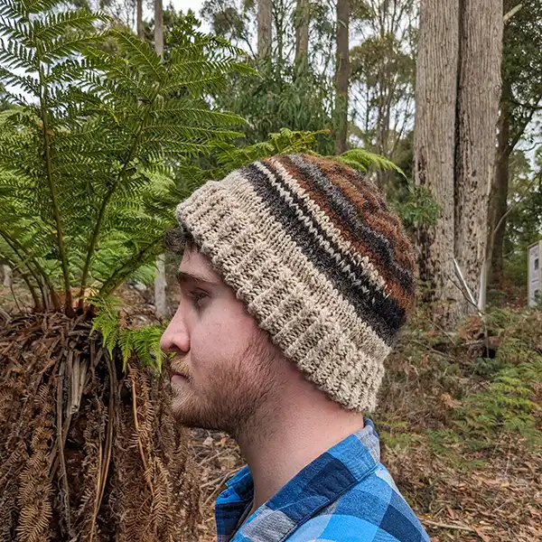 A young man is facing to the side. He is wearing a hand-knitted beanie made from a mixture of coloured fleeces from gey to dark brown